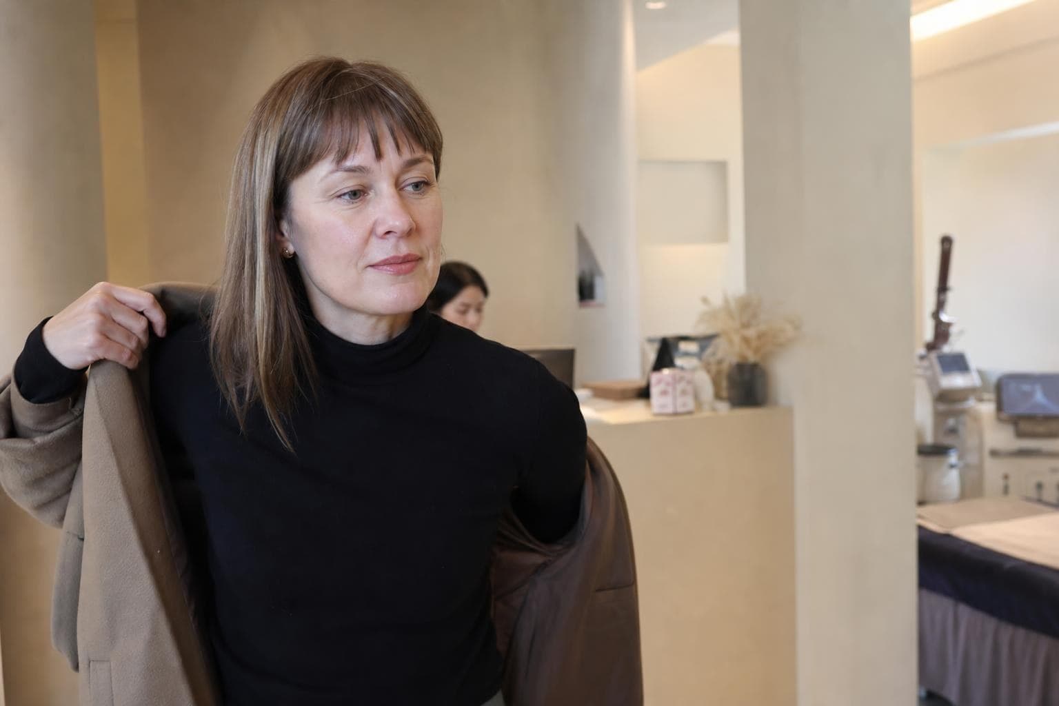 A woman in her thirties touching her jawline in a mirror, with soft daylight, before a laser consultation in Seoul